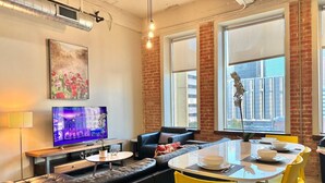 A modern living room with a black leather sofa, a dining table with yellow chairs, a flat-screen TV, and large windows with a city view. - 2 BR | 2 BT | Downtown | Free Valet Parking | # 6 (Dallas)
