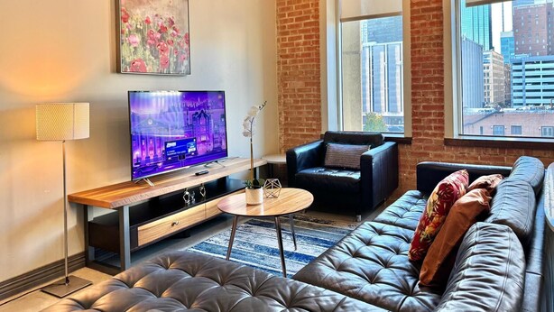A modern living room with a leather sofa, a flat-screen TV, a coffee table, and a large window with a city view. - 2 BR | 2 BT | Downtown | Free Valet Parking | # 6 (Dallas)