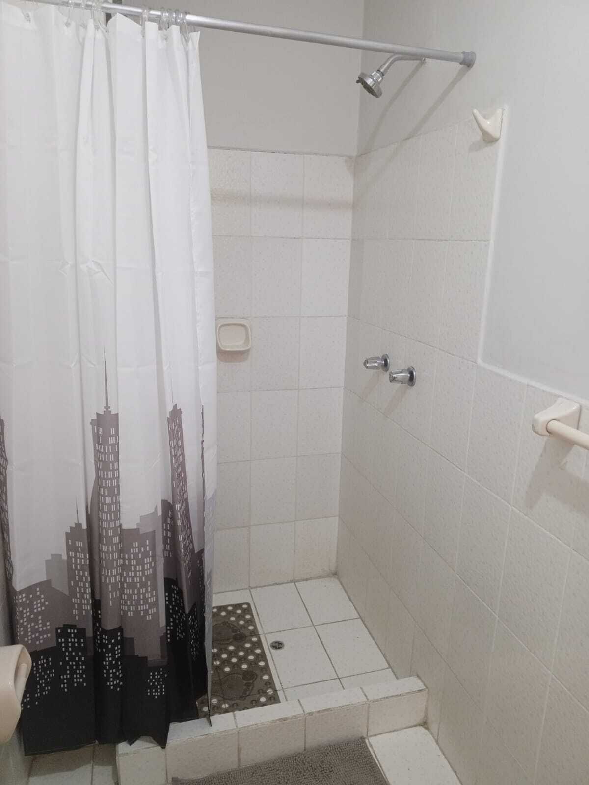 Un espace douche avec un mur recouvert de carreaux blancs, une pomme de douche, une barre d’appui et un rideau de douche à motifs noirs et blancs.