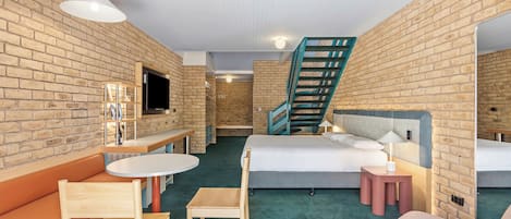 A hotel room with a brick wall, a bed, a desk, a chair, a sofa, and a staircase.