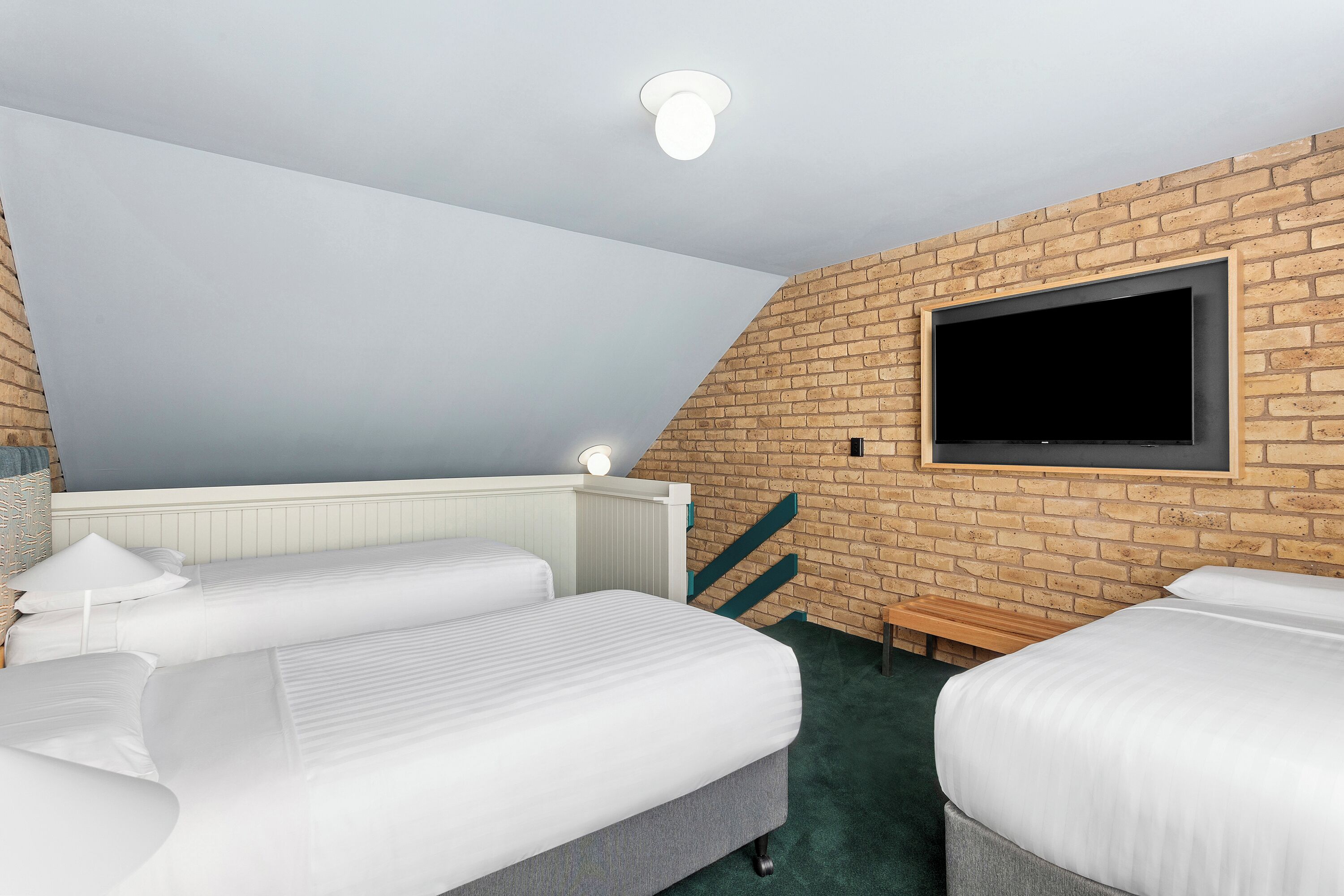 A hotel room with two beds, a TV mounted on a brick wall, a white radiator, and a ceiling light.