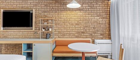 A hotel room with a brick wall, a flat-screen TV, a wooden desk, a small kitchenette, a round table, a chair, and a bed.