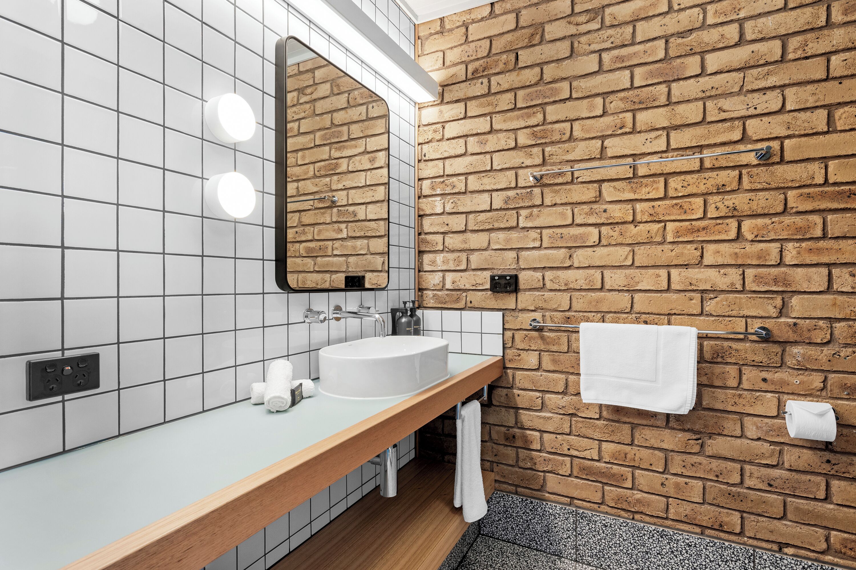 A bathroom with a white sink, a mirror, and a towel rack against a brick wall.