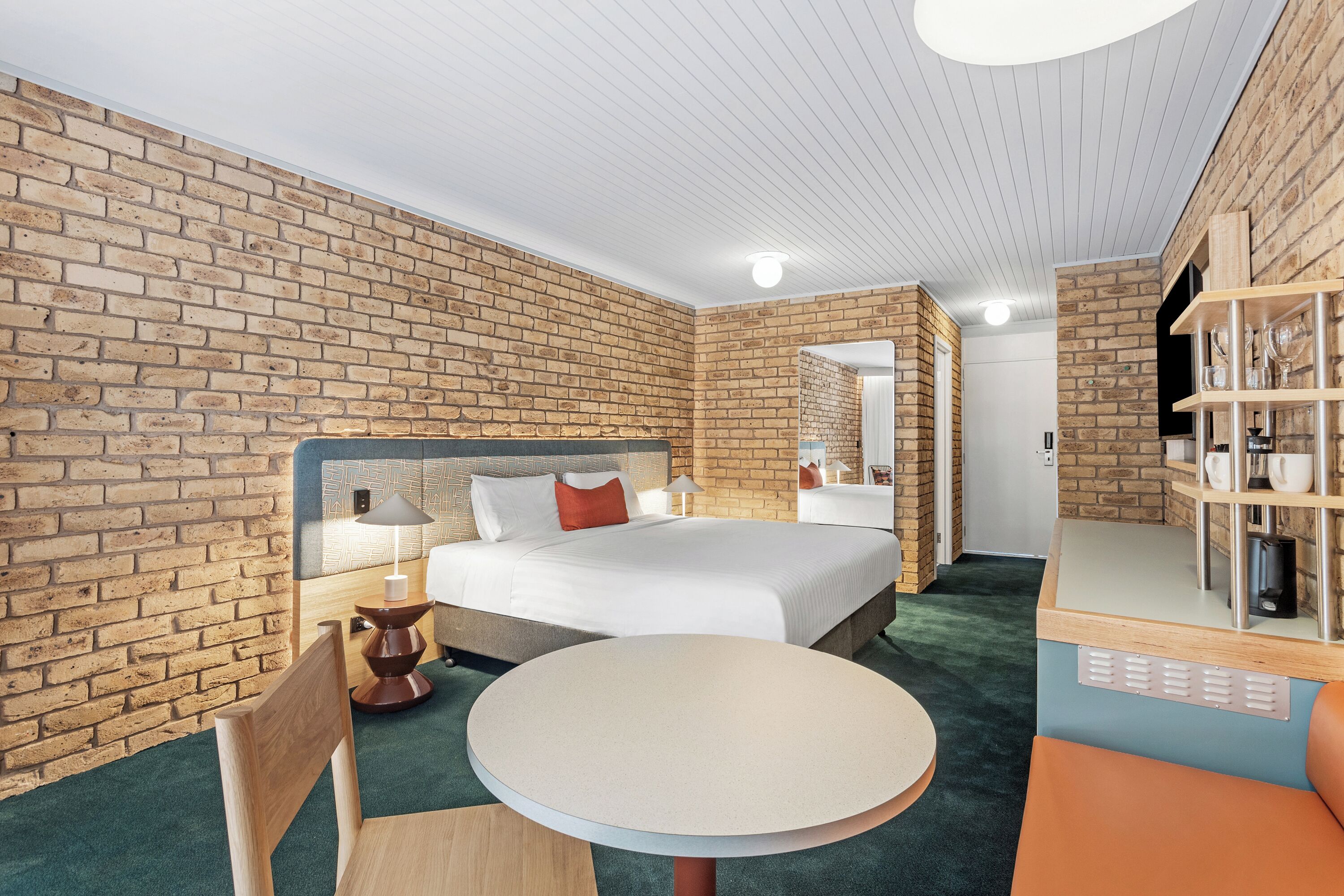 Une chambre d’hôtel avec un mur en briques, un lit avec une literie blanche, une table ronde et un coin salon avec un fauteuil.
