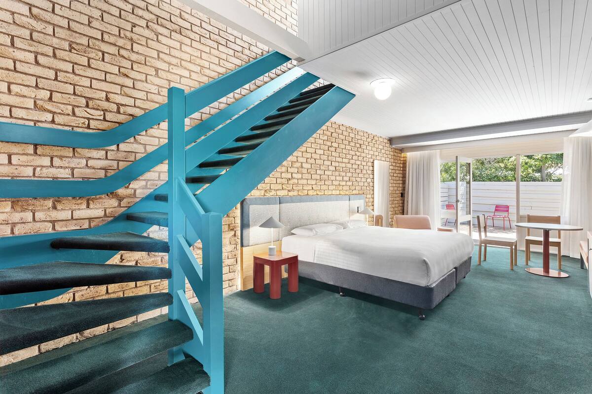 Une chambre d’hôtel moderne avec un mur en briques, un escalier bleu, un lit, une petite table et une chaise.