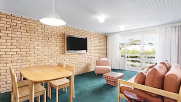 Een kamer met een bakstenen muur, een flatscreen televisie, een houten tafel met stoelen, een roze fauteuil en een bank met kussens.