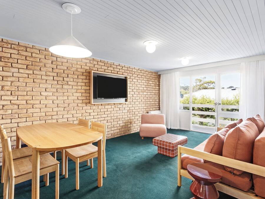 Een kamer met een bakstenen muur, een flatscreen televisie, een houten tafel met stoelen, een roze fauteuil en een bank met kussens.