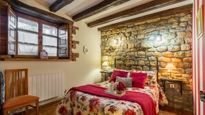 Un dormitorio con pared de piedra, cama de madera, ventana con cortinas, una silla y una mesita de noche con lámpara.