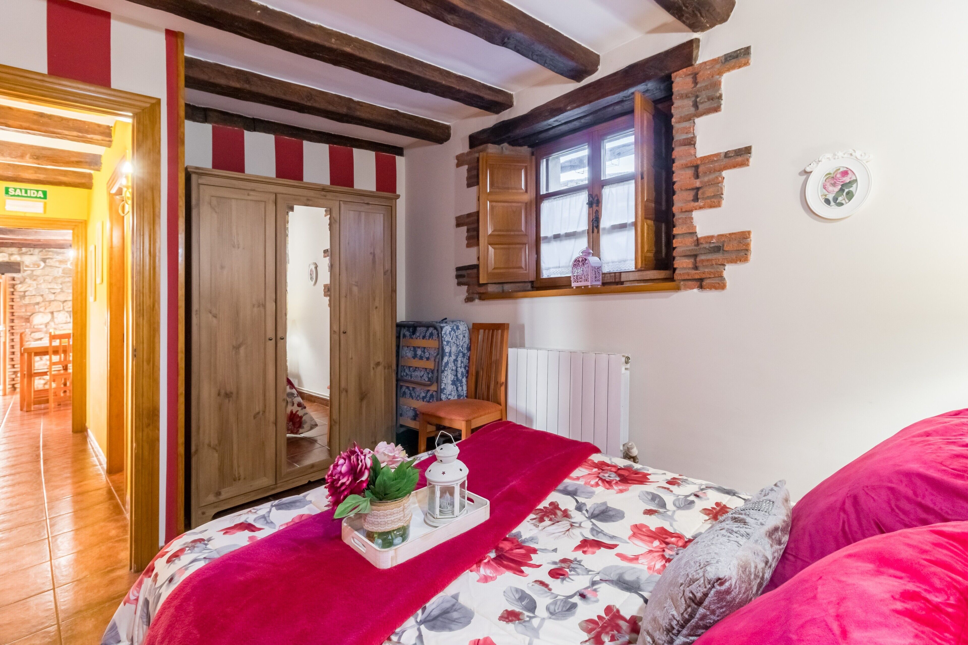Un dormitorio con un armario de madera, una cama con ropa de cama floral, un plaid rojo, una mesita con un farol y una ventana con vistas al exterior.