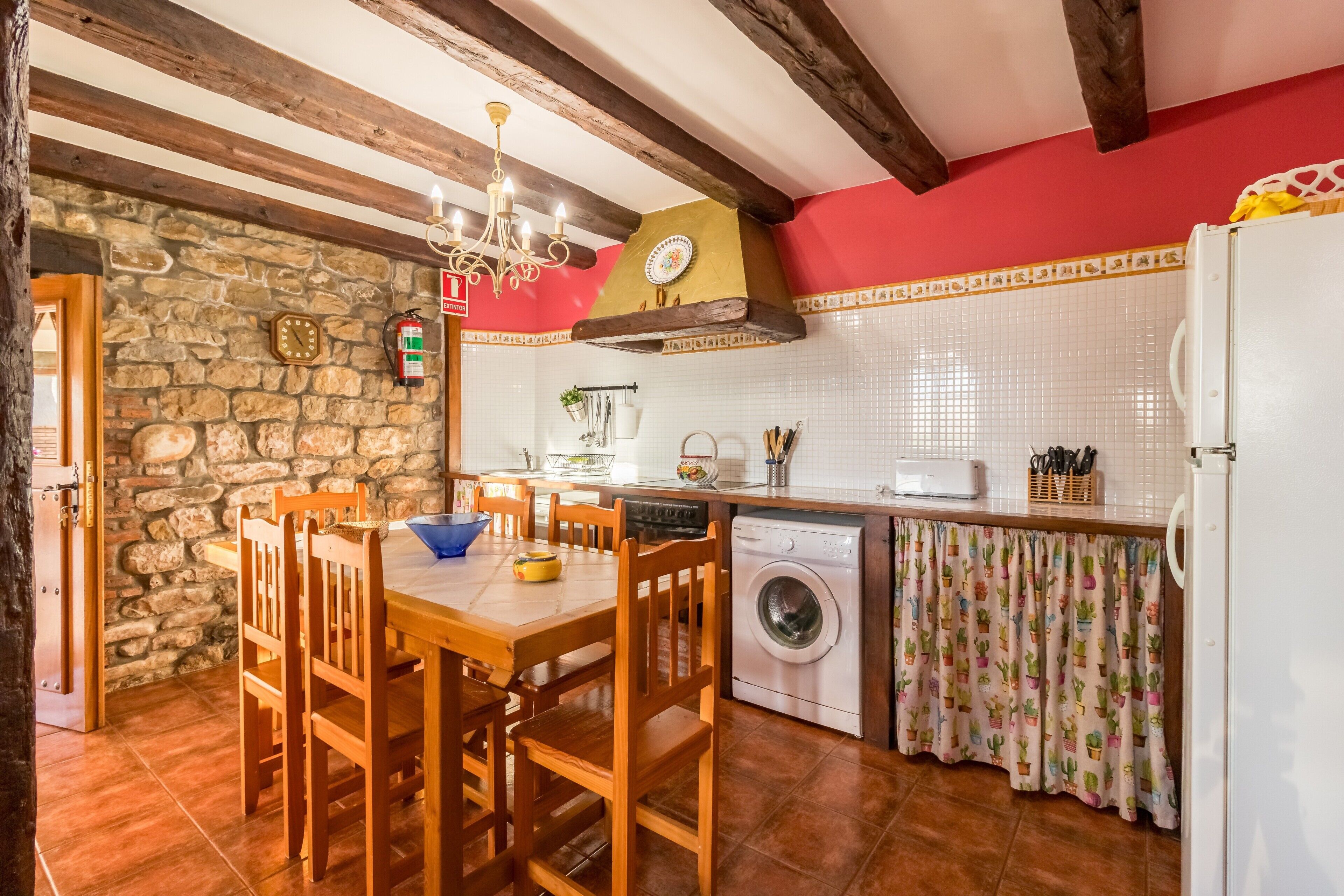 A kitchen with a stone wall, wooden ceiling beams, a washing machine, and a dining table with chairs.