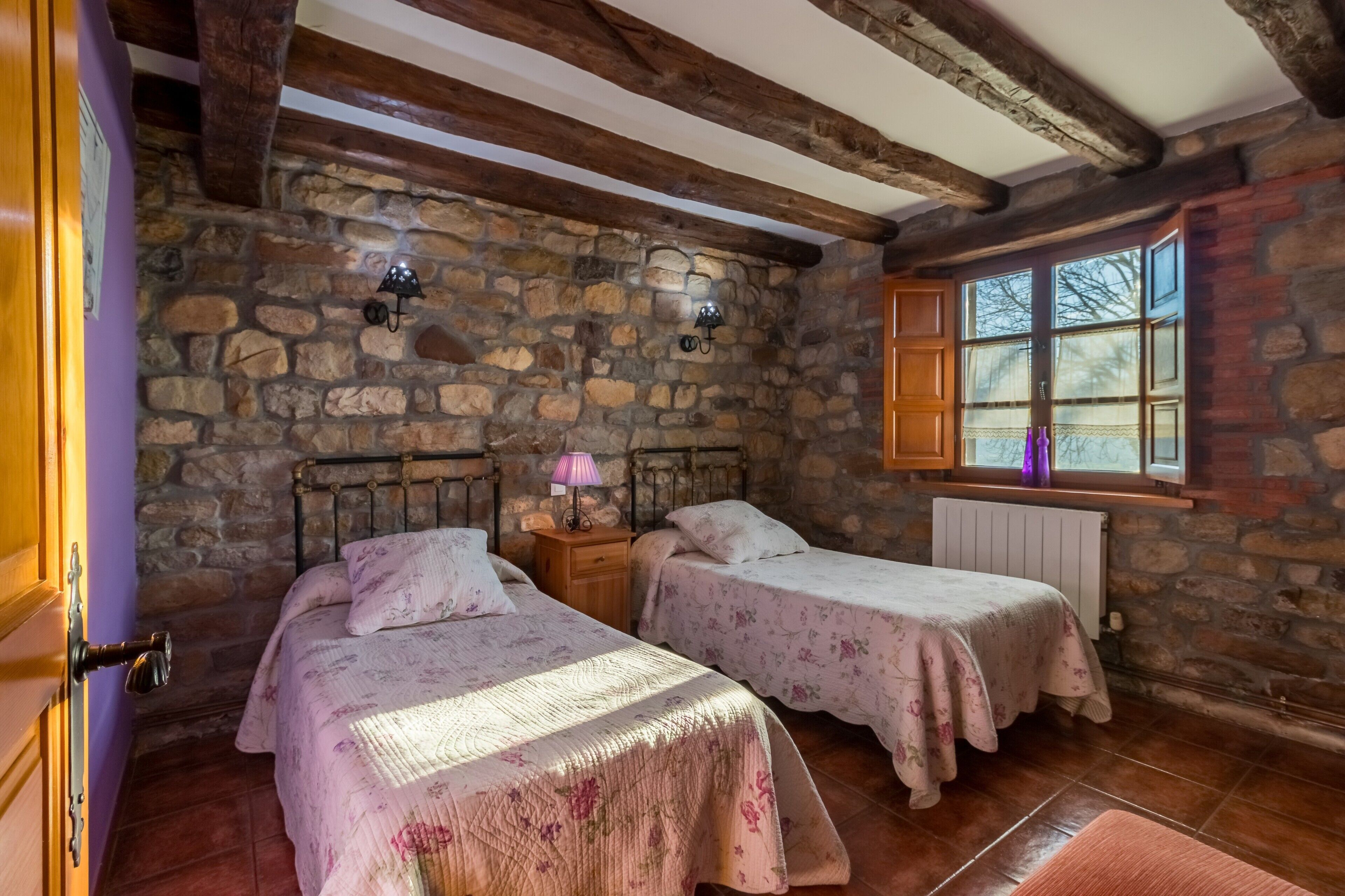 Una habitación rústica con paredes de piedra, vigas de madera, dos camas con ropa de cama floral, una ventana con vistas y una mesita de noche con lámpara.