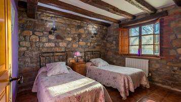 Una habitación rústica con paredes de piedra, vigas de madera, dos camas con ropa de cama floral, una ventana con vistas y una mesita de noche con lámpara.