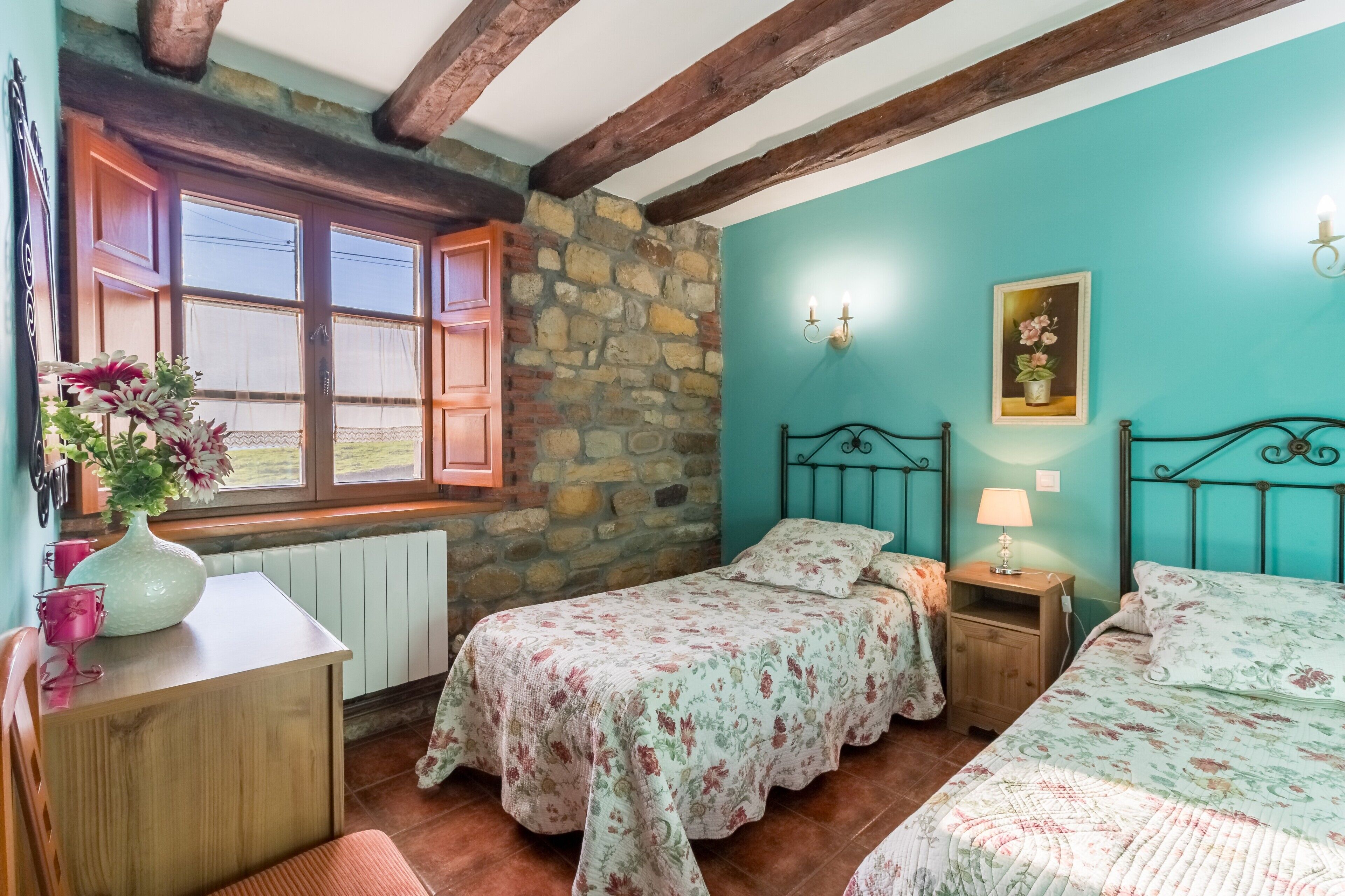 A bedroom with two beds, a stone wall, wooden beams, a window with shutters, a vase with flowers, and a lamp on a bedside table.