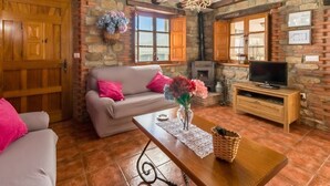 Una sala de estar acogedora con pared de piedra, vigas de madera, chimenea, televisor y un sofá con cojines rosa.