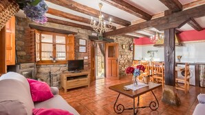 Un interior rústico con vigas de madera a la vista, pared de piedra, televisor, comedor y cocina.
