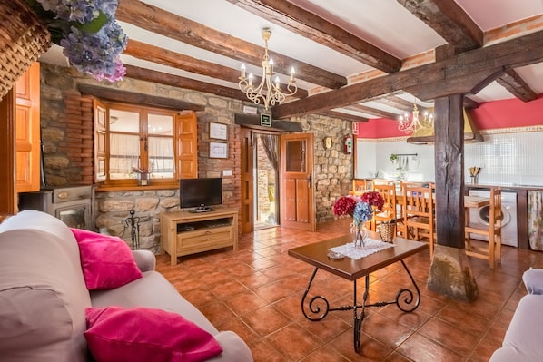 Un intérieur rustique avec des poutres apparentes, un mur en pierre, un téléviseur, un coin repas et une cuisine.