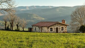 A stone house with a red roof, a chimney, and a picnic table in a grassy field with mountains in the background.