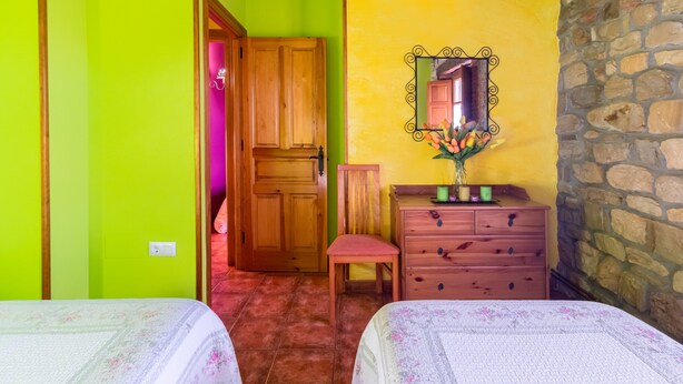 Une chambre avec deux lits, une armoire en bois, un miroir orné de motifs floraux et un mur en pierre.
