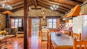 A rustic kitchen with stone walls, wooden beams, a tiled floor, a dining table, chairs, a refrigerator, a microwave, and a television.