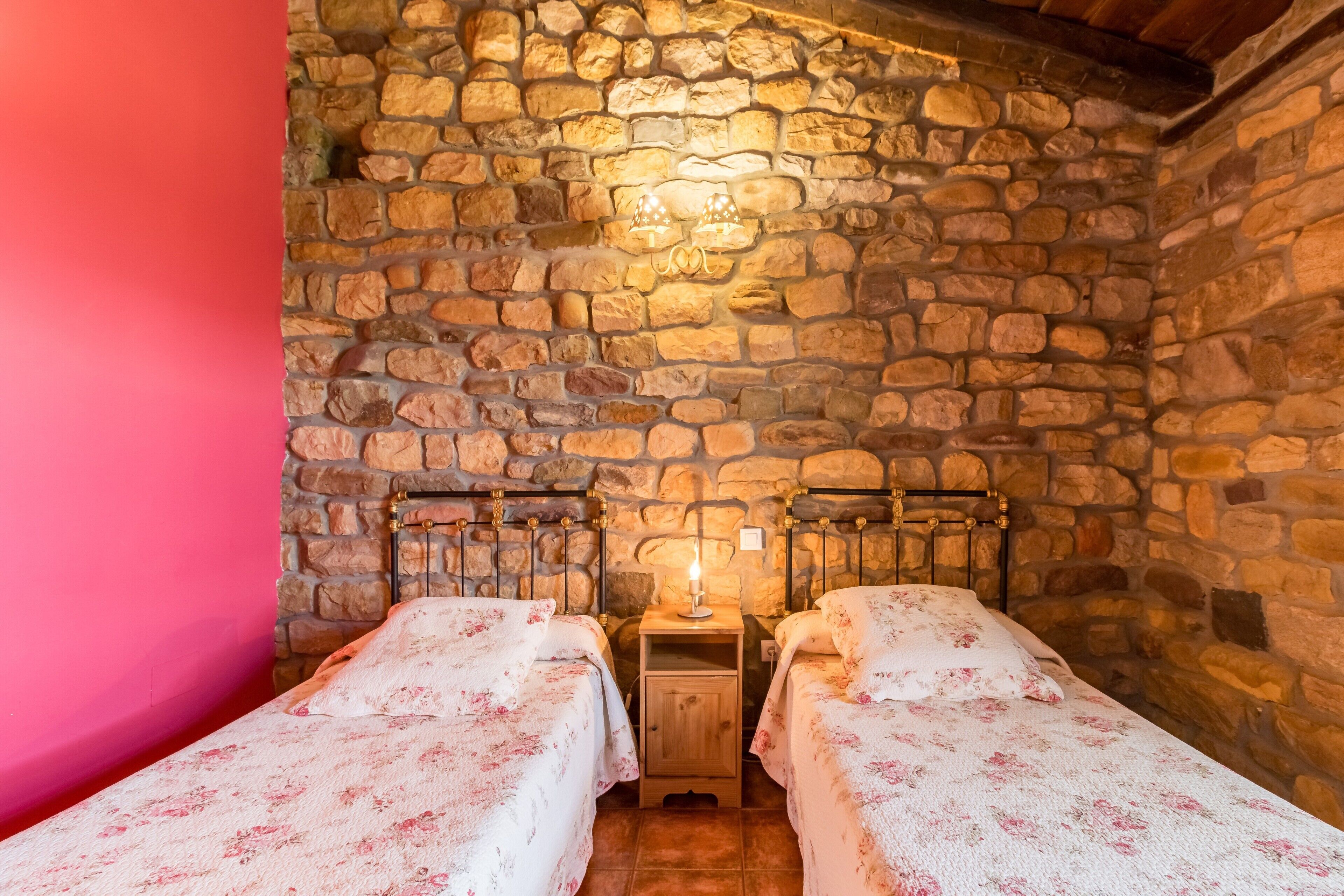 A room with stone walls, two beds with floral bedding, a nightstand with a lamp, and a wooden ceiling.