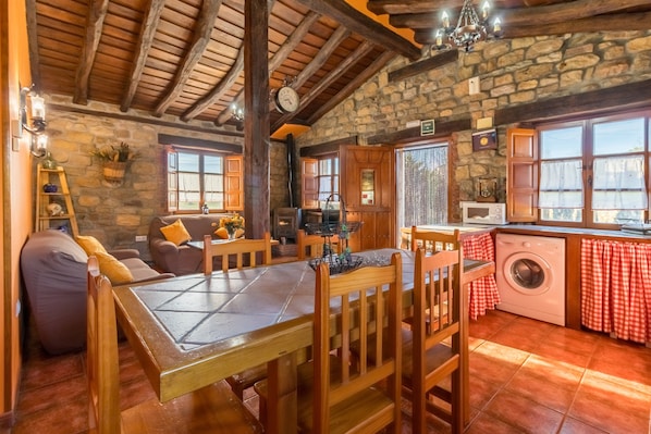 A rustic interior with a stone wall, wooden beams, a dining table, chairs, a sofa, a kitchen area with a washing machine, and a window with curtains.