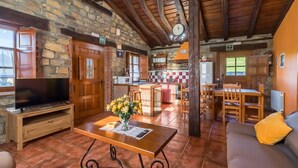 A rustic interior with stone walls, wooden beams, a television, a dining area, and a kitchen.