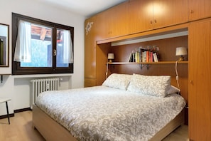 Une chambre à coucher avec un lit, une armoire en bois, une étagère avec des livres et une lampe, ainsi qu’une fenêtre avec des rideaux.