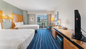 Habitación de hotel con dos camas, un escritorio con computadora, una silla y vista a la ciudad a través de amplias ventanas.