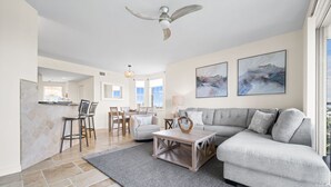 A spacious living room with a grey sofa, a coffee table, and a dining area with chairs and a table. There are two large windows providing natural light and a ceiling fan.