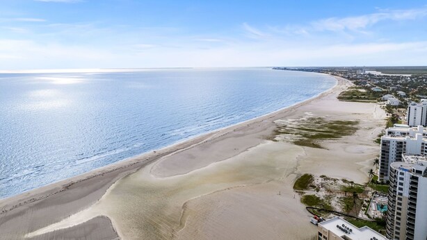 An aerial view of a beach with a clear sky, a sandy shoreline, and adjacent high-rise buildings.