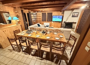 Un comedor con una mesa de madera dispuesta para cuatro personas, rodeada de sillas, con un televisor y estanterías de fondo.