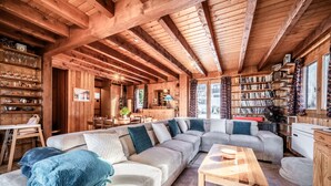 A cozy wooden interior with a large sectional sofa, a wooden table, and a bookshelf filled with books. - Chalet 'Des Charmes' with Mountain Views, Private Terrace, and Wi-Fi (Thollon-les-Mémises)