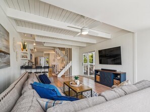 A spacious living room with a large flat-screen TV, a ceiling fan, and a staircase leading to an outdoor area. - Expansive Coastal Retreat, 50 Yards from the Beach (Bethany Beach)