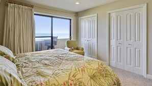 A bedroom with a large bed, a chair, a window with a view, and a set of white doors.