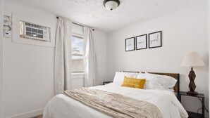 Ein Schlafzimmer mit einem Bett, einem Nachttisch, einer Lampe, einem Fenster mit Vorhängen und drei gerahmten Bildern an der Wand.