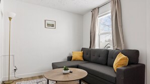 Ein Wohnzimmer mit grauem Sofa, zwei senfgelben Kissen, einem runden Couchtisch mit Pflanze, einer Stehlampe und einem gerahmten Bild an der Wand.