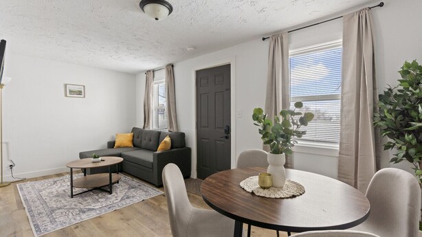 Ein modernes Wohnzimmer mit grauem Sofa, einem kleinen runden Tisch und einem Esstisch mit Stühlen. Auf dem Esstisch steht eine Pflanze und es gibt ein Fenster mit Vorhängen.