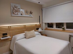 A bedroom with a wooden headboard, a bed with white bedding, a bedside table with a lamp, and a window with a blind.