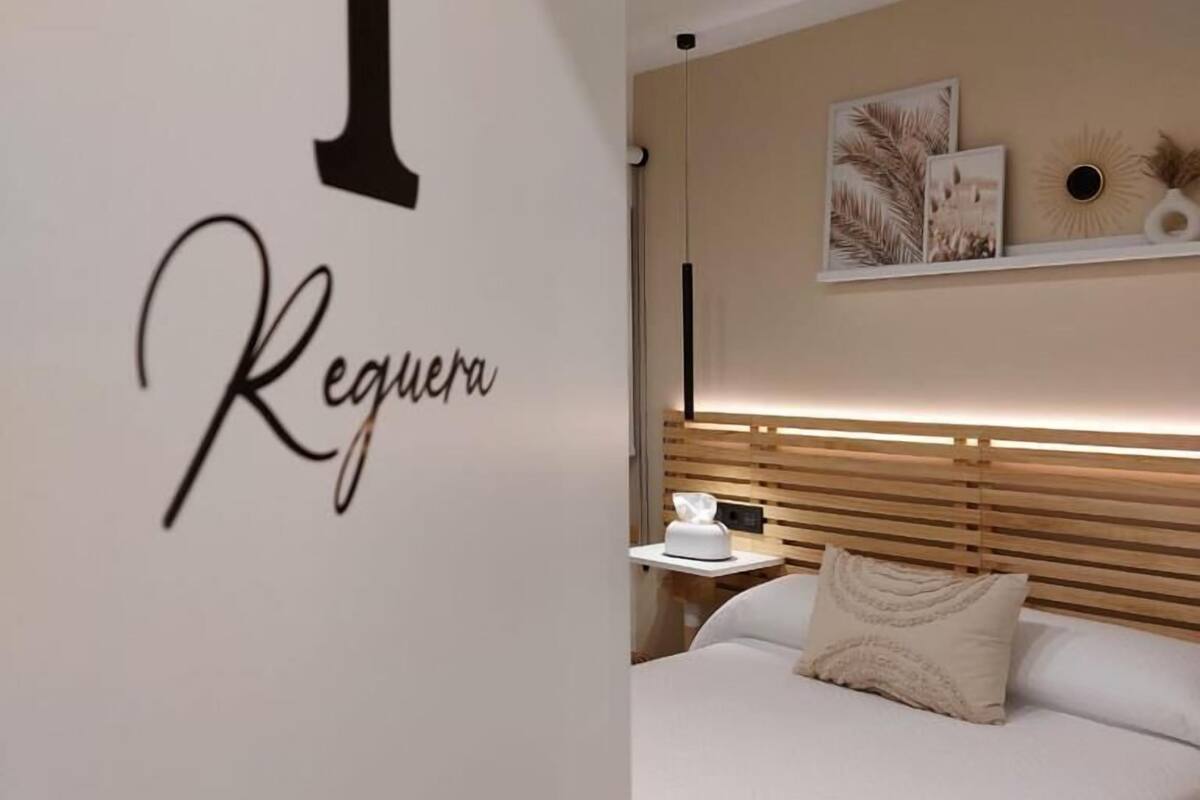 Une chambre d’hôtel avec un grand numéro « 1 » et le mot « Reguera » sur le mur, un lit avec du linge de lit blanc, une table de chevet avec une lampe et une étagère avec des objets décoratifs.