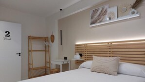 Een slaapkamer met een bed, nachtkastje, houten hoofdbord, wandplank met decoratieve objecten en een deur met een nummer.