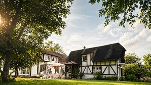 Une maison traditionnelle à colombages, dotée d’une pelouse bien entretenue et d’un grand arbre qui offre de l’ombre.