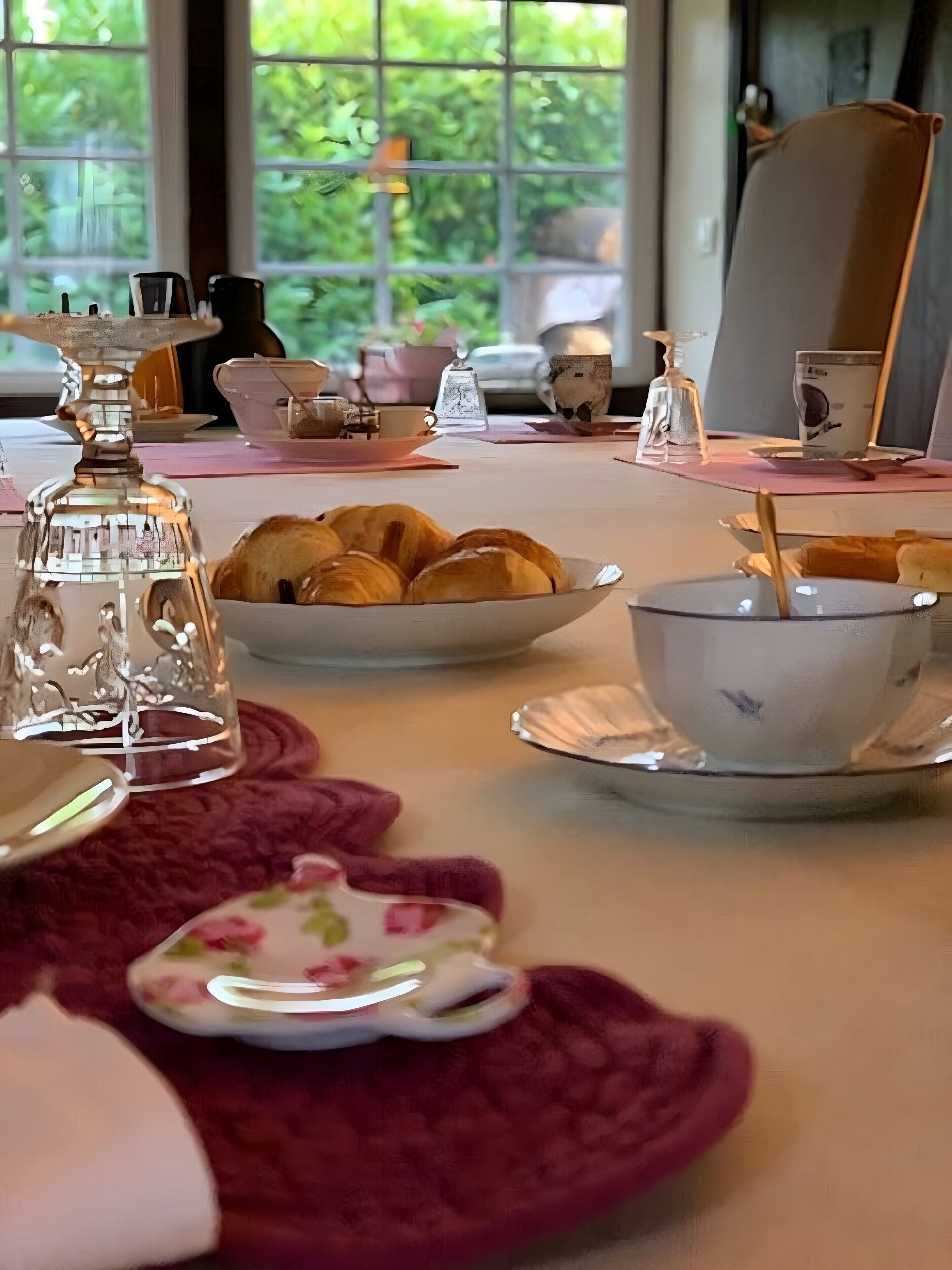 Une table à manger ornée d’une carafe en verre transparent, d’un panier de croissants, d’une assiette à motifs floraux et d’une théière en argent.