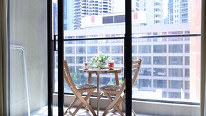 A balcony with a table and chairs, a potted plant, and a window with a view of tall buildings. - Modern Studio + Balcony + Car Park - Darling Harbour (Sydney)