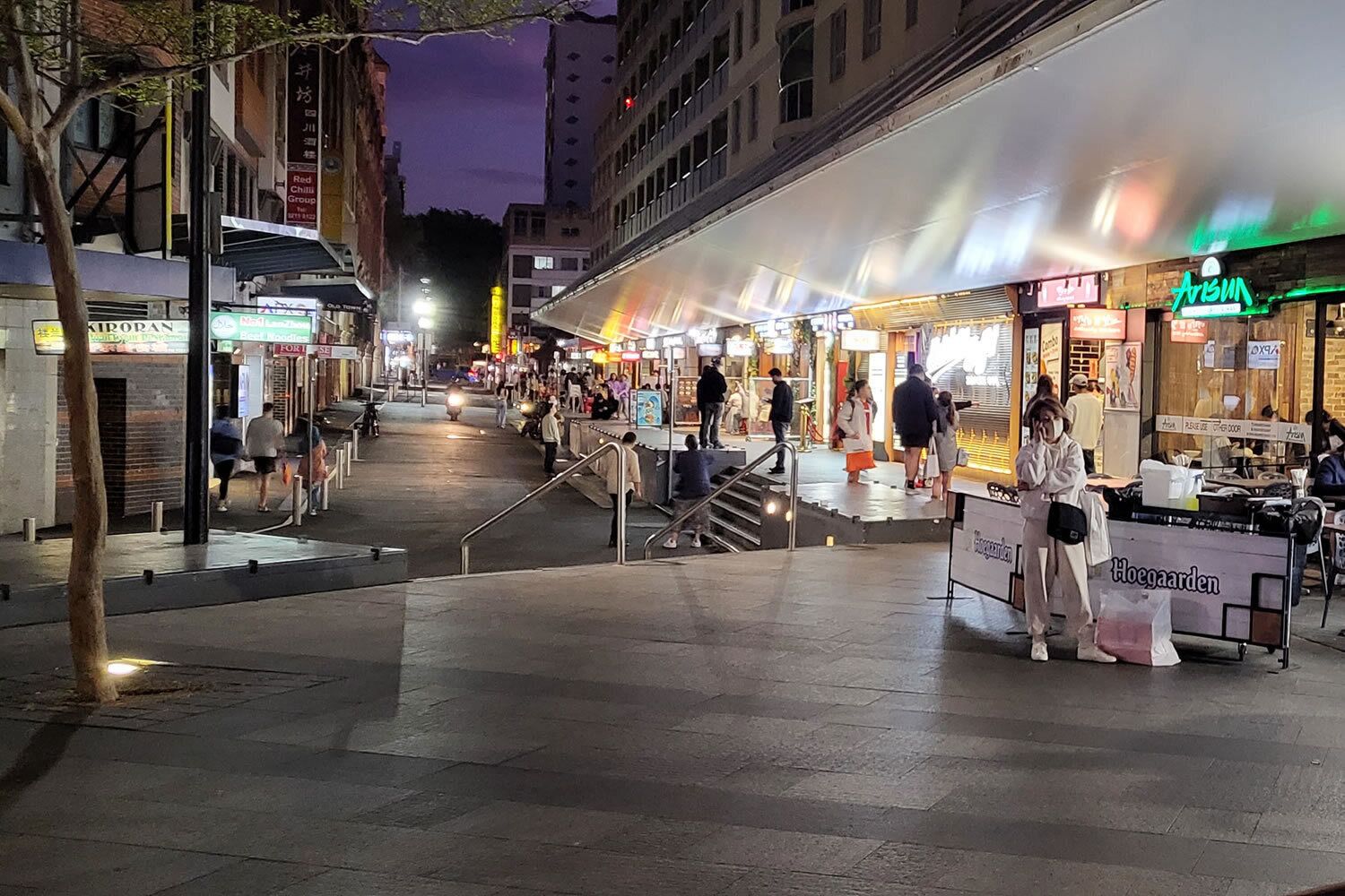 A nighttime street scene with outdoor cafes and shops.