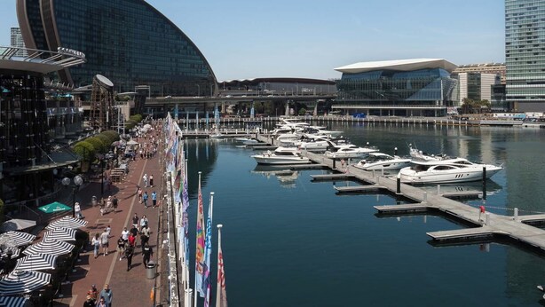 A marina with boats docked, a modern building with a 'W' logo, and a pedestrian walkway with umbrellas. - Modern Studio + Balcony + Car Park - Darling Harbour (Sydney)