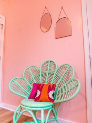Una silla color turquesa con un cojín llamativo frente a una pared rosa, junto con dos espejos de pared.