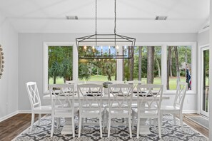 A dining area with a white table and chairs, a geometric chandelier, and large windows with a view of trees and a lawn. - RBC Heritage Views From Your Deck, 4BR Villa Steps to Harbour Town (Hilton Head Island)