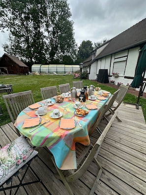 Eine Holzterrasse mit gedecktem Tisch, umgeben von Stühlen und einem Gewächshaus im Hintergrund.