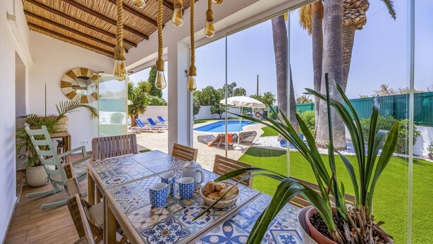 A dining area with a table set for four, a pool, and a view of palm trees and a fence.