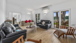 A spacious living area with a large sofa, a smaller sofa, and a dining table. There are two doors leading to other rooms, and a large sliding glass door to the outside.